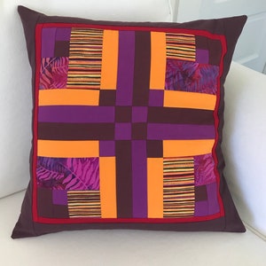 May include: A square pillow with a geometric pattern in shades of orange, purple, and brown. The pillow is made of fabric with a quilted design.