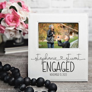 May include: White picture frame with a stitched border. The frame has a photo of a couple getting engaged in a park. The text on the frame reads "Stephanie & Stuart ENGAGED NOVEMBER 13, 2023".
