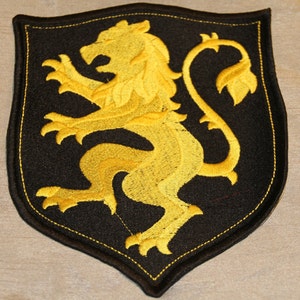 Könnte beinhalten: Ein schwarzer gestickter Aufnäher in Form eines Schildes mit einem goldenen Löwen in der Mitte.