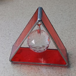 May include: A red stained glass triangle with a clear, faceted crystal ball suspended inside. The glass is framed with silver-colored metal. The crystal ball hangs from a thin wire, adding a touch of elegance to this decorative piece.
