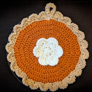 May include: A handmade crochet trivet in the shape of a pie. The center is orange, with a white flower in the middle. The crust is tan, and the trivet has a loop for hanging. The trivet is on a black surface.