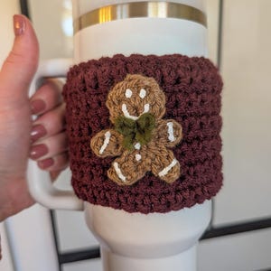 May include: A white travel mug with a burgundy crochet cozy featuring a gingerbread man design. The cozy is hand-crocheted with a gingerbread man applique in brown, white, and green. The mug has a white handle and a gold band.