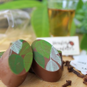 May include: Four oval-shaped chocolate truffles with a green leaf design. The truffles are arranged on a wooden surface with cinnamon sticks and cloves.