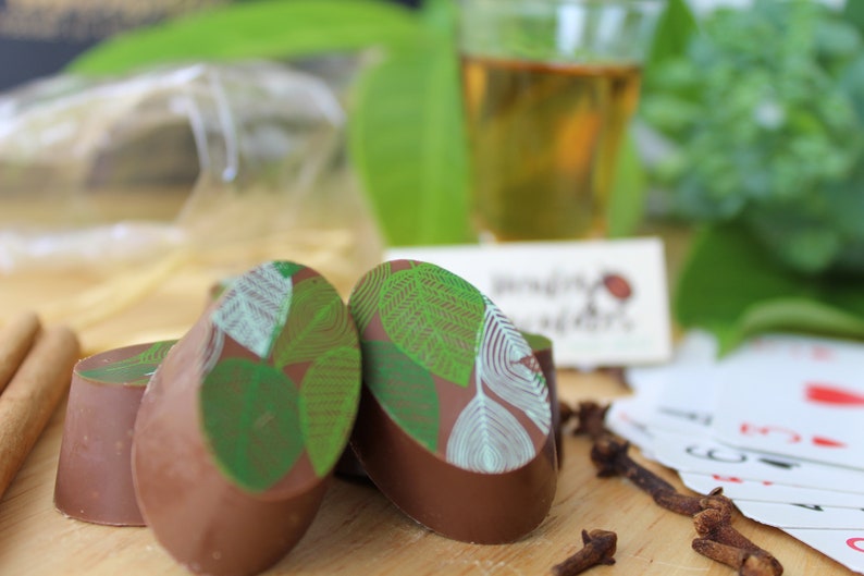 May include: Four oval-shaped chocolate truffles with a green leaf design. The truffles are arranged on a wooden surface with cinnamon sticks and cloves.
