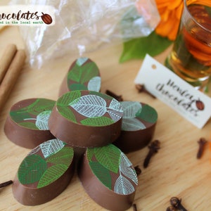 May include: A close-up of a stack of oval-shaped chocolates with a green and white leaf design. The chocolates are arranged on a wooden surface with cinnamon sticks, cloves, and a small glass of amber liquid.