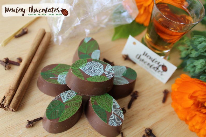 May include: A close-up of a stack of oval-shaped chocolates with a green and white leaf design. The chocolates are arranged on a wooden surface with cinnamon sticks, cloves, and a small glass of amber liquid.