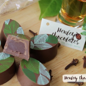 May include: A close-up of a wooden surface with a variety of chocolate truffles. The truffles are oval-shaped and have a green and brown leaf design. The text "Henley Chocolates rooted in the local earth" is visible in the image.