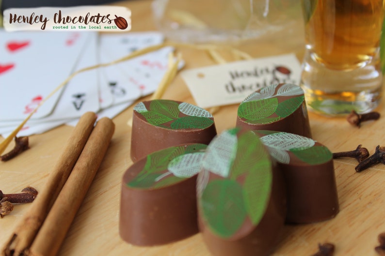 May include: A close-up of five chocolate truffles with a green leaf design. The truffles are arranged on a wooden surface with cinnamon sticks, cloves, and a glass of amber liquid.