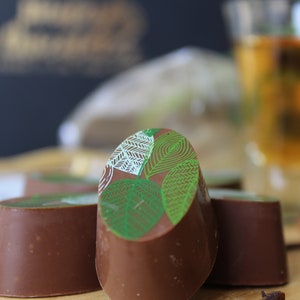 May include: Four chocolate truffles with a green leaf design on top. The truffles are arranged in a row on a wooden surface. There is a glass of amber liquid and cinnamon sticks in the background.