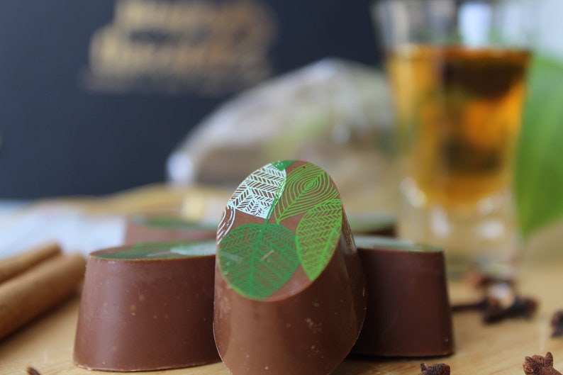May include: Four chocolate truffles with a green leaf design on top. The truffles are arranged in a row on a wooden surface. There is a glass of amber liquid and cinnamon sticks in the background.