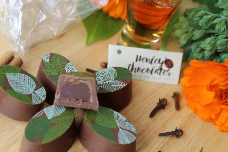 May include: A close-up of six oval-shaped chocolate truffles with a green and white leaf design. The truffles are arranged on a wooden surface with a small tag that reads "Henley Chocolates".