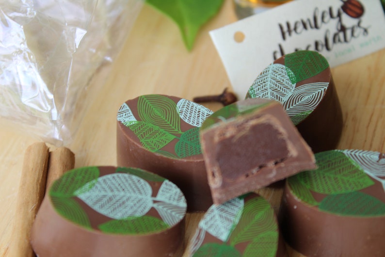 May include: Assortment of chocolate truffles with a green and white leaf design. The truffles are arranged on a wooden surface. Henley Chocolates is printed on a white tag.