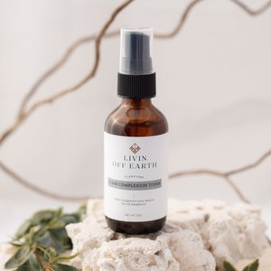 Könnte beinhalten: Eine braune Glasflasche mit schwarzem Sprühkopf und einem weißen Etikett mit der Aufschrift "Livin Off Earth Clarifying Clear Complexion Toner". Die Flasche steht auf einem weißen Stein.