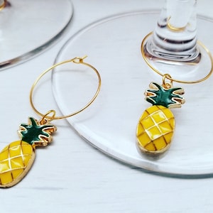 May include: Gold-colored wine glass charms with a yellow pineapple design. The pineapple charms have green tops and are attached to gold hoops. The charms are on a clear glass surface.
