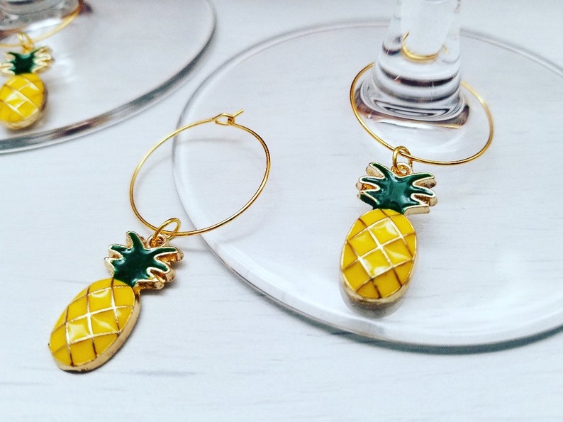 May include: Gold-colored wine glass charms with a yellow pineapple design. The pineapple charms have green tops and are attached to gold hoops. The charms are on a clear glass surface.