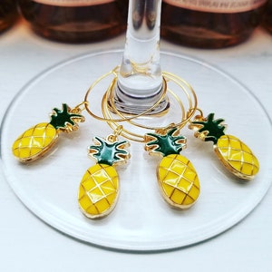 May include: Set of five gold-colored wine glass charms with yellow pineapple pendants and green tops. The charms are hanging on a wine glass. The pineapple charms have a gold border.