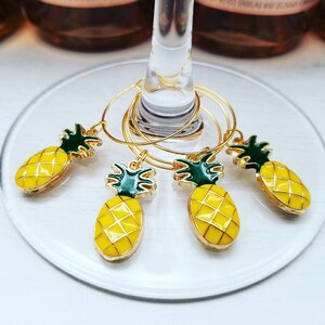 May include: Set of four gold-colored wine glass charms with yellow pineapple pendants and green tops. The charms are hanging on a wine glass. The pineapple charms are yellow with a gold outline.