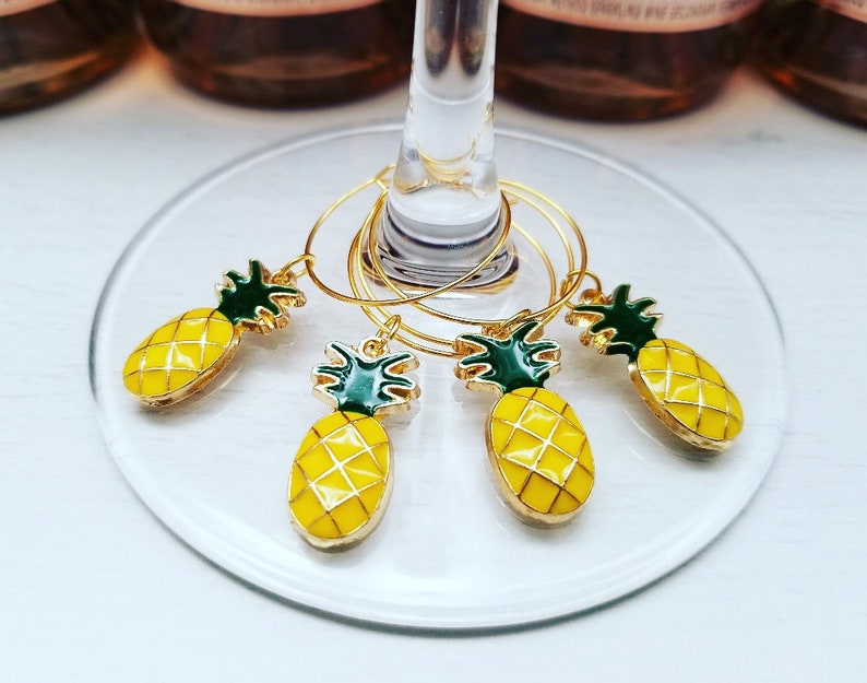 May include: Set of four gold-colored wine glass charms with yellow pineapple pendants and green tops. The charms are hanging on a wine glass. The pineapple charms are yellow with a gold outline.