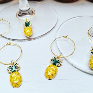 May include: Gold-colored wine charms with a yellow pineapple design and green accents. The pineapple charms are attached to a gold hoop, designed to hang on the stem of a wine glass. The charms are on a white surface.