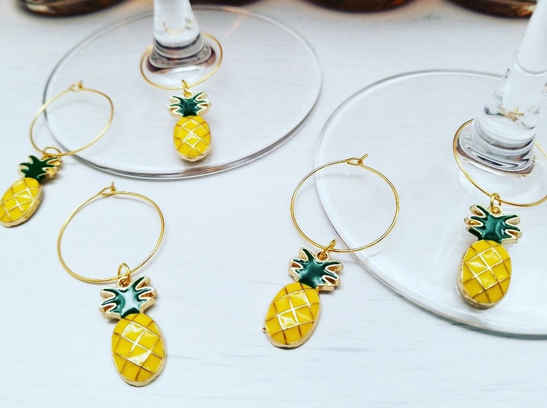 May include: Gold-colored wine charms with a yellow pineapple design and green accents. The pineapple charms are attached to a gold hoop, designed to hang on the stem of a wine glass. The charms are on a white surface.