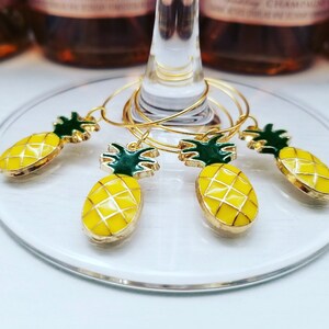 May include: Set of five gold-toned wine glass charms with yellow pineapple pendants and green tops. The charms are on a clear glass surface, with bottles of rose wine in the background.