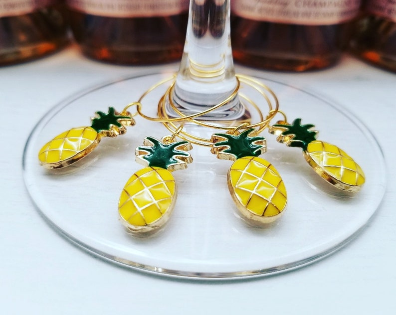 May include: Set of five gold-toned wine glass charms with yellow pineapple pendants and green tops. The charms are on a clear glass surface, with bottles of rose wine in the background.