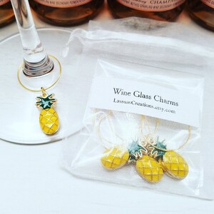 May include: Gold-toned wine glass charms with yellow pineapple pendants and green tops. The charms are displayed on a wine glass and in a small, clear bag. The bag has a label that reads "Wine Glass Charms" and the Etsy shop name.