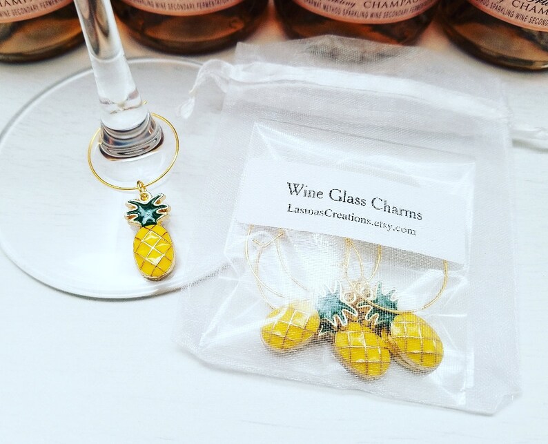 May include: Gold-toned wine glass charms with yellow pineapple pendants and green tops. The charms are displayed on a wine glass and in a small, clear bag. The bag has a label that reads "Wine Glass Charms" and the Etsy shop name.