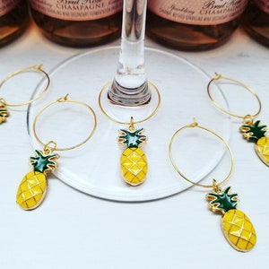 May include: Gold-colored wine glass charms with yellow pineapple pendants and green accents. The charms are hanging on a wine glass and are set against a background of champagne bottles.