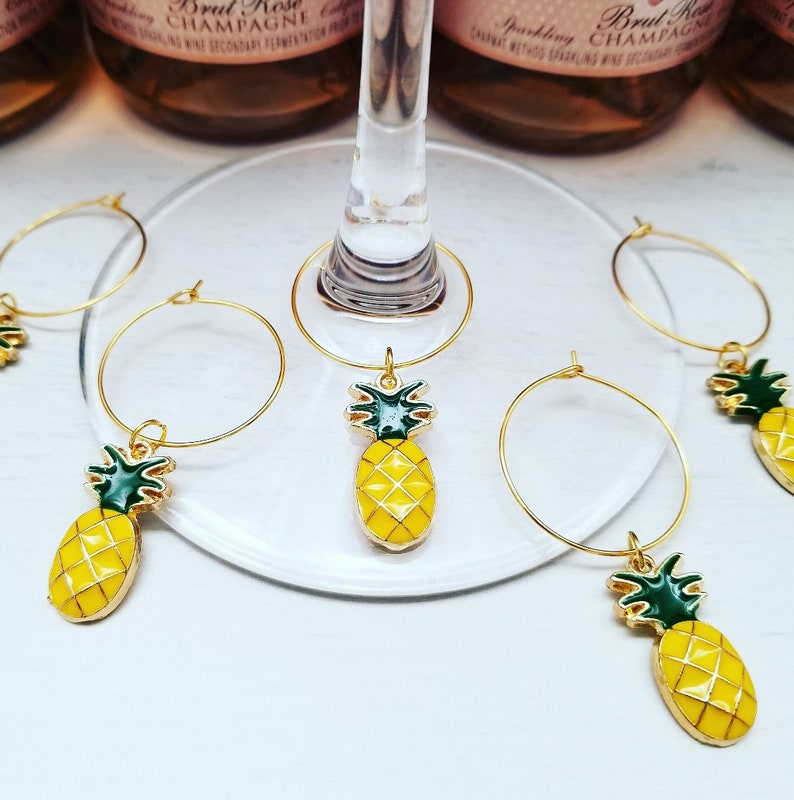 May include: Gold-colored wine glass charms with yellow pineapple pendants and green accents. The charms are hanging on a wine glass and are set against a background of champagne bottles.