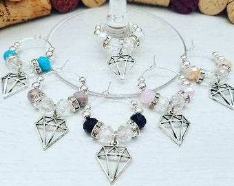 May include: A collection of wine glass charms with silver-colored diamond pendants. Each charm features a different color of beads, including turquoise, pink, and clear, with silver accents. The charms are designed to hang on the stem of a wine glass.