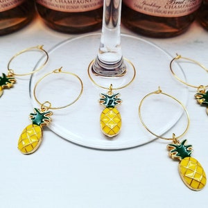 May include: Gold-toned wine glass charms with yellow pineapple pendants and green accents. The charms are arranged on a clear glass surface, with bottles of sparkling wine in the background. The pineapple charms add a tropical touch.