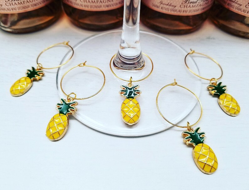 May include: Gold-toned wine glass charms with yellow pineapple pendants and green accents. The charms are arranged on a clear glass surface, with bottles of sparkling wine in the background. The pineapple charms add a tropical touch.