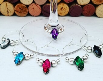 May include: A collection of wine glass charms with silver hoops and colorful gemstone pendants. The charms feature clear, blue, pink, green, and black gemstones, and are accented with small silver beads. The background includes wine corks.