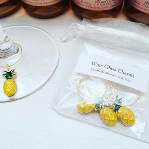 May include: Gold-toned wine glass charms with yellow pineapple pendants and green leaves. The charms are displayed on a wine glass and in a small white bag. Text on the bag reads "Wine Glass Charms" and a website address.