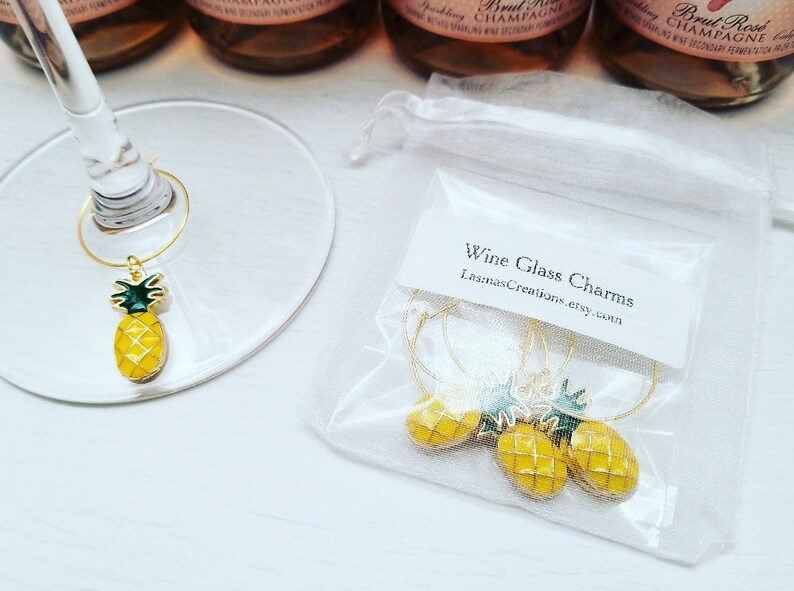 May include: Gold-toned wine glass charms with yellow pineapple pendants and green leaves. The charms are displayed on a wine glass and in a small white bag. Text on the bag reads "Wine Glass Charms" and a website address.