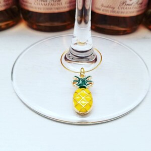 May include: A gold-colored wine charm with a yellow and green pineapple design. The charm is hanging from a wine glass. The background includes bottles of sparkling wine.