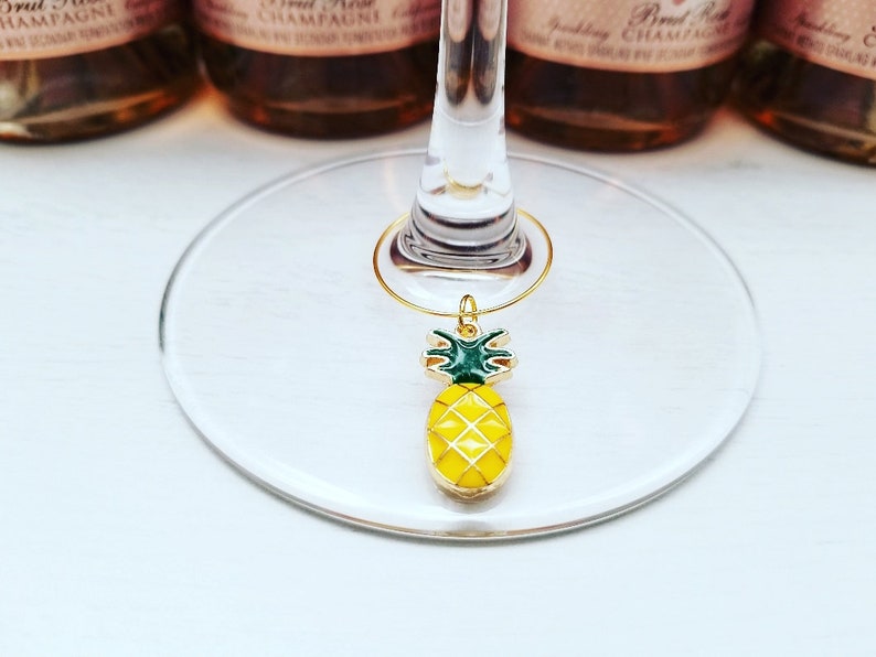 May include: A gold-colored wine charm with a yellow and green pineapple design. The charm is hanging from a wine glass. The background includes bottles of sparkling wine.