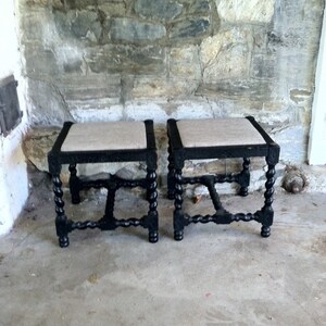 May include: Two black wooden stools with a woven fabric seat. The stools have a turned leg design and are in a rustic setting.