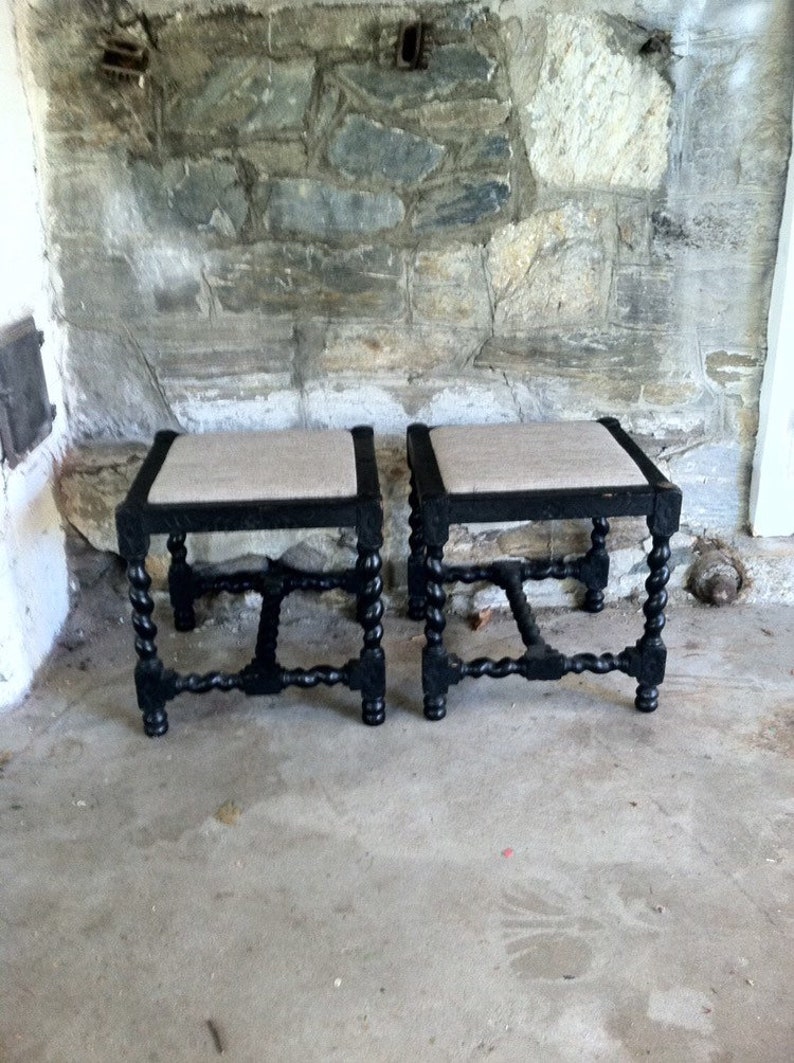 May include: Two black wooden stools with a woven fabric seat. The stools have a turned leg design and are in a rustic setting.