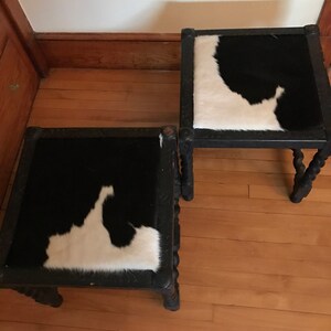May include: Two black wooden stools with a cowhide seat. The stools have a carved design on the legs and a black and white cowhide seat.
