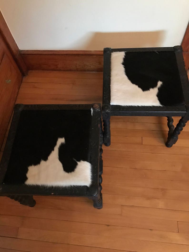 May include: Two black wooden stools with a cowhide seat. The stools have a carved design on the legs and a black and white cowhide seat.