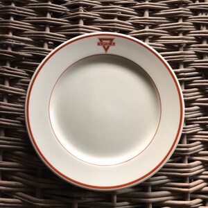 May include: A white ceramic plate with a red rim and a red YMCA logo in the center. The plate is on a woven wicker surface.