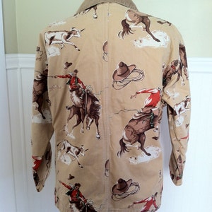 Pu&ograve; includere: Giacca beige a tema cowboy con colletto in velluto a coste. La giacca presenta un motivo ripetuto di cowboy che cavalcano cavalli selvaggi, cavalli e bovini. Il design include cappelli e corde da cowboy marroni. La giacca &egrave; esposta su un manichino nero.