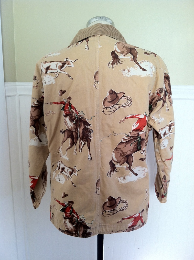 Pu&ograve; includere: Giacca beige a tema cowboy con colletto in velluto a coste. La giacca presenta un motivo ripetuto di cowboy che cavalcano cavalli selvaggi, cavalli e bovini. Il design include cappelli e corde da cowboy marroni. La giacca &egrave; esposta su un manichino nero.