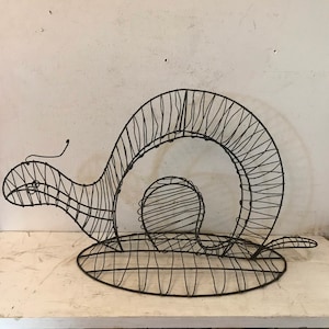 May include: A wire sculpture of a snail, with a rounded shell and a long, thin body. The sculpture is made of black wire and is designed to be a decorative piece.