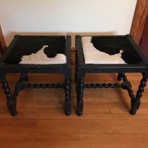 May include: A pair of black wooden stools with twisted legs and cowhide seats. The cowhide is black and white.