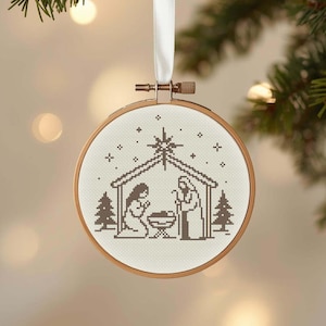 Può includere: Un ornamento natalizio rotondo con un disegno a punto croce raffigurante la Natività. L'ornamento è incastonato in un telaio da ricamo in legno e appeso a un nastro bianco. Il disegno presenta Maria, Giuseppe, il bambino Gesù e una stella.