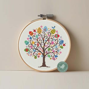 Pode incluir: Um bastidor de bordado em ponto cruz com um desenho de árvore adornado com ovos de Páscoa coloridos. Os ovos apresentam vários padrões e cores, incluindo vermelho, azul, amarelo e verde. O desenho está sobre um fundo branco, emoldurado por um aro de madeira.