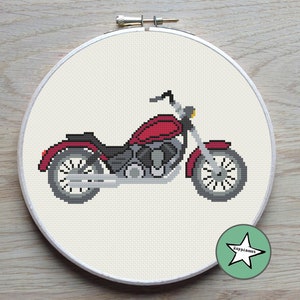 May include: A cross-stitch embroidery of a red motorcycle with gray and black details, set in a white hoop. The motorcycle has a classic design with a fuel tank, seat, and wheels. The background is a light beige fabric.